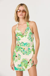 A woman is wearing a sleeveless, halter-neck dress with a colorful tropical floral pattern in shades of green, pink, and blue on a light cream background. The dress has a fitted bodice and a slightly loose, short skirt.